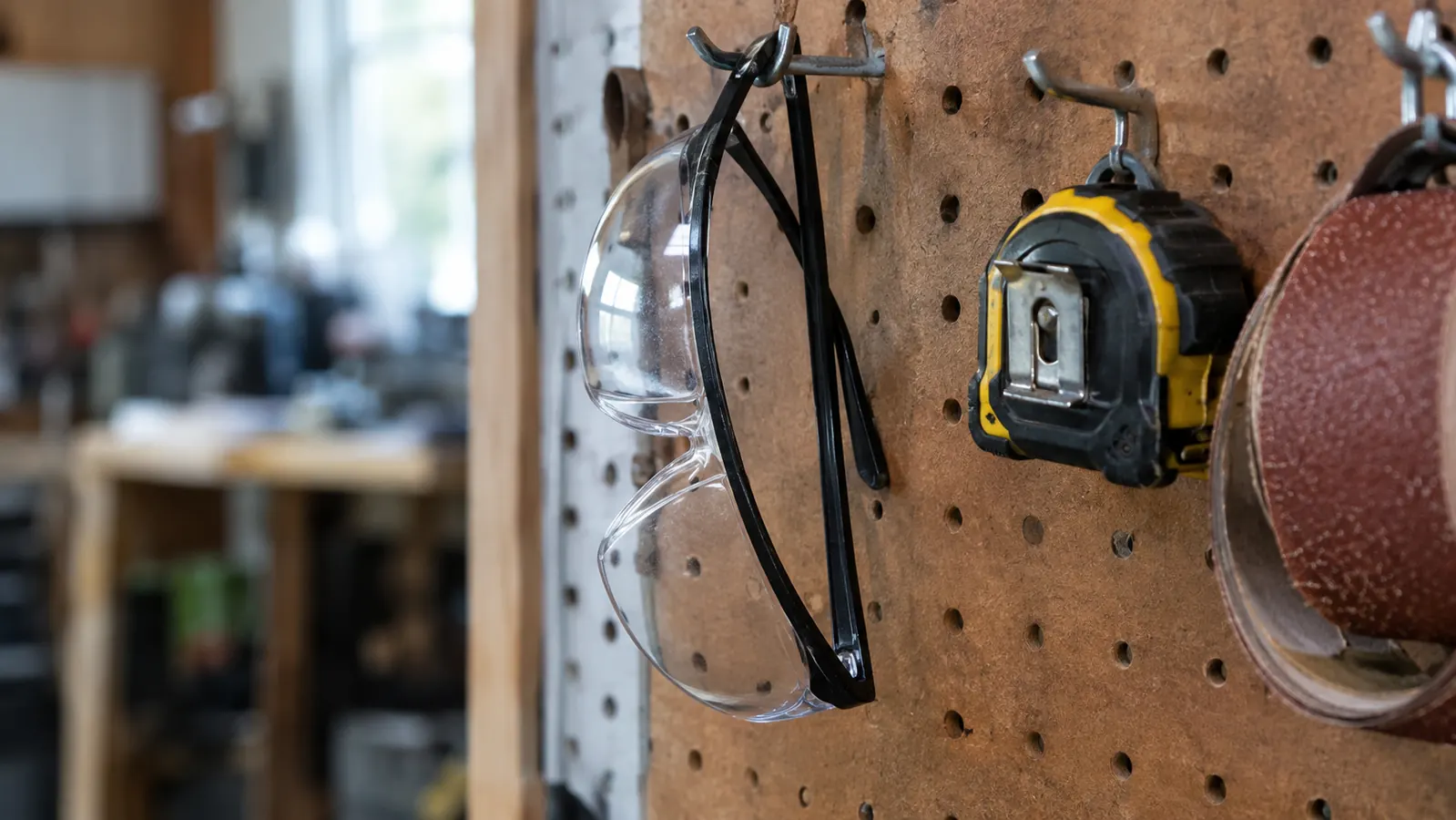 Safety glasses hanging from a pegboard hook beside a tape measure and a roll of sandpaper in a workshop